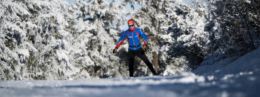 Zu sehen ist jemand, der Ski im Schnee fährt. Er trägt ein blaues Oberteil und eine schwarze Hose. Außerdem trägt er noch eine rote Kopfbedeckung und eine Sonnenbrille.