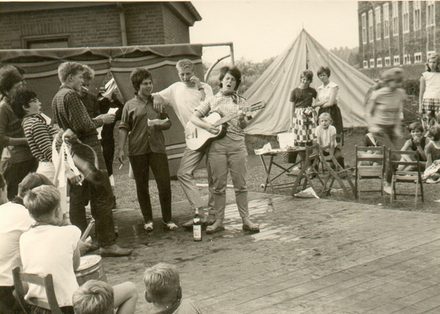 Bild: Mehrere Kinder stehen im Kreis. Eines der Kinder spielt Gitarre. Im Hintergrund ist ein Zelt zu sehen. 