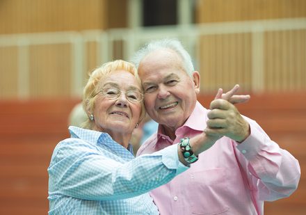 Bild: Älteres Pärchen, Mann und Frau, beim Tanzen. Beide schauen in die Kamera. 