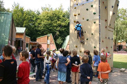 Bild: Viele Kinder stehen um eine Kletterwand. An der Kletterwand klettert ein Kind nach oben. 