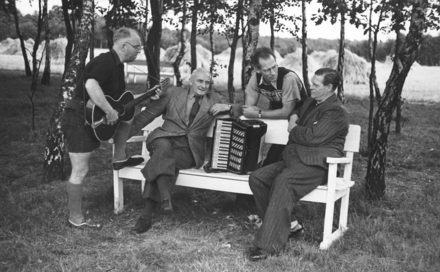 Bild: Zwei Männer sitzen auf einer Bank. Zwischen ihnen steht ein Akkordeon. Vorne steht ein Mann mit einem Bein auf der Bank und spielt Gitarre. Hinter der Bank steht ein weiterer Mann. 