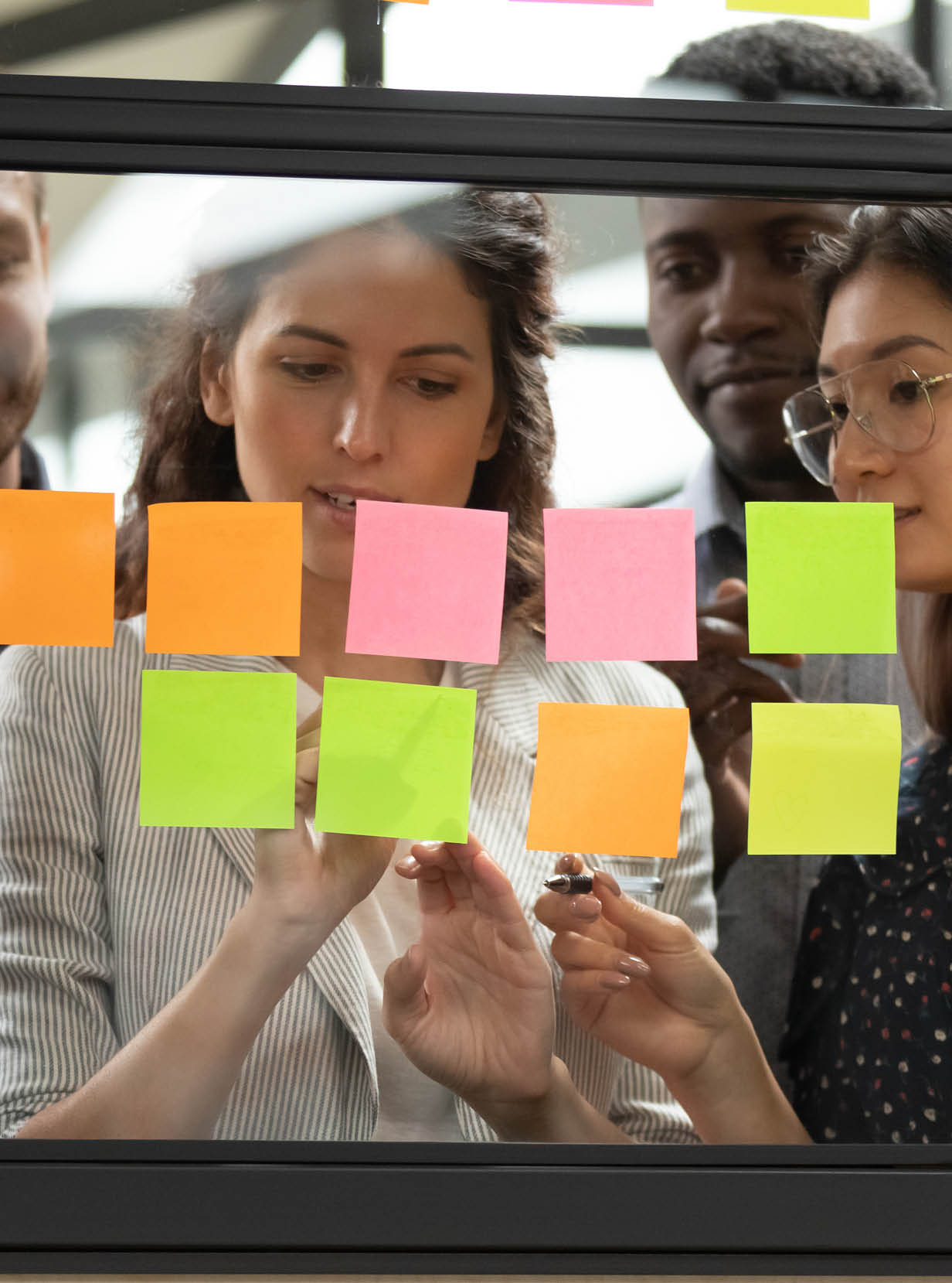 Focused young diverse teammates working with caucasian team leader on IT startup project kanban organization process, discussing details and tasks, writing notes on colorful paper stickers at office.